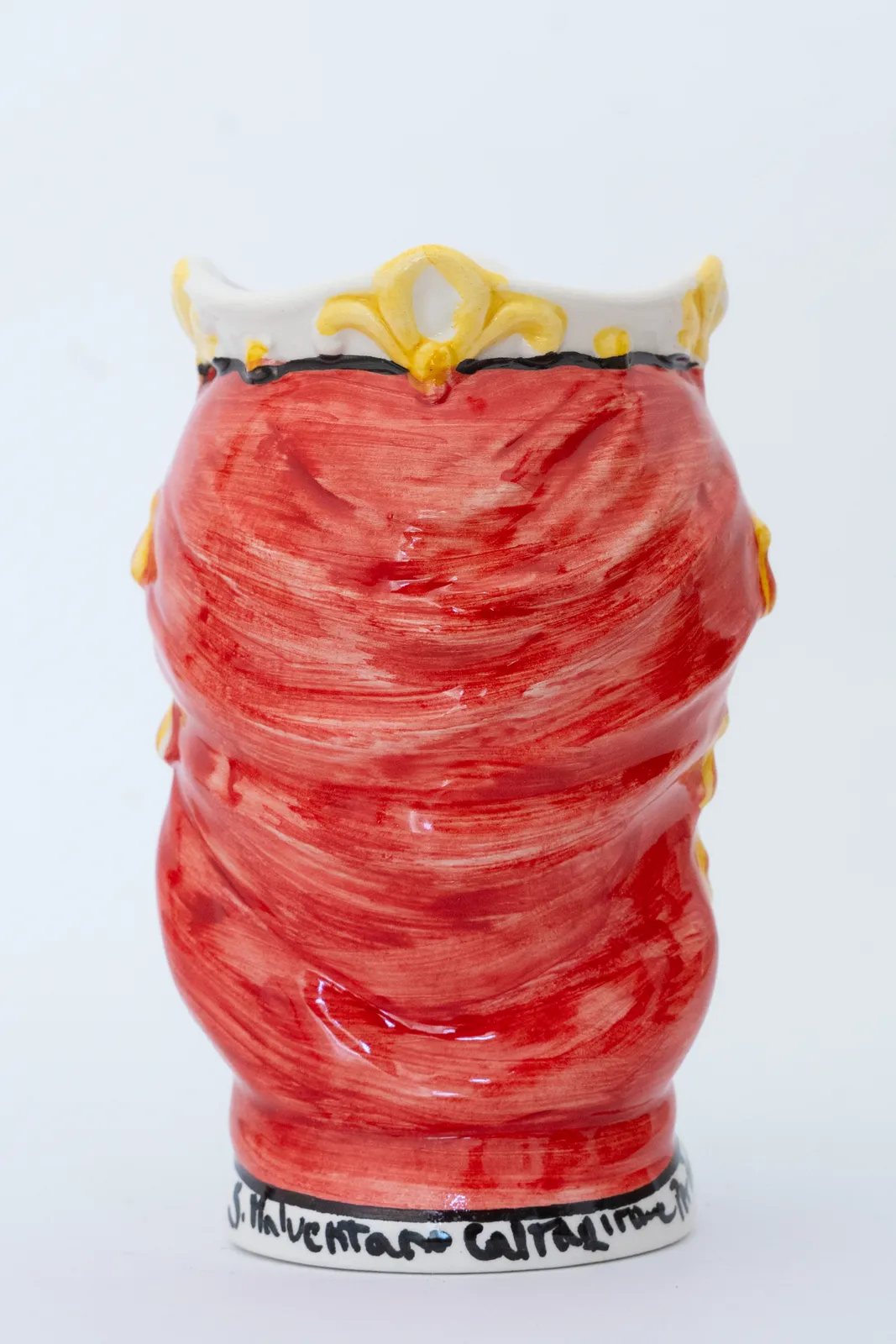Un vaso decorativo in ceramica con un design rosso e strutturato che ricorda un drappeggio in tessuto. La parte superiore è ornata da accenti ornamentali gialli e una scritta nera è visibile alla base. Lo sfondo è bianco semplice.